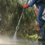 Person pressure-washing sidewalk with a few shrubs in the foreground of the photo